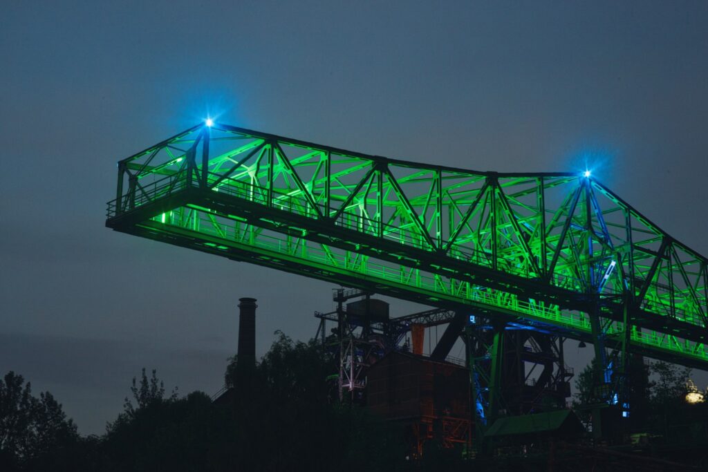 Grün beleuchteter Kran, genannt Krokodil, im Landschaftspark Duisburg-Nord vor dem blauen Abendhimmel