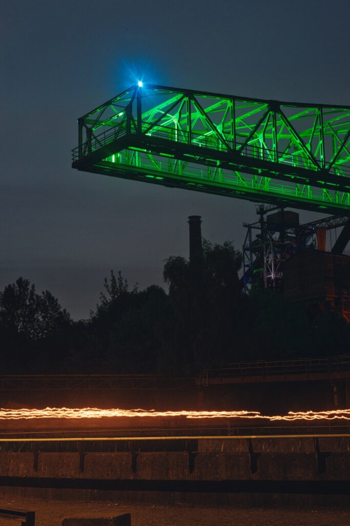 Lichtspuren unter einer grün beleuchteten Stahlkonstruktion im Landschaftspark Duisburg-Nord bei Nacht